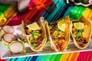 A white plate topped with three tacos and radishes on a colorful table cloth.