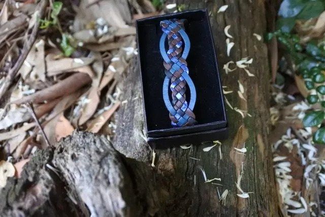Braided blue and brown bracelet in a black box, resting on a tree trunk outdoors.