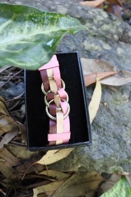 Bracelet in a black box, with pink, brown, and tan woven straps, and gold rings. Set on a rock with leaves.