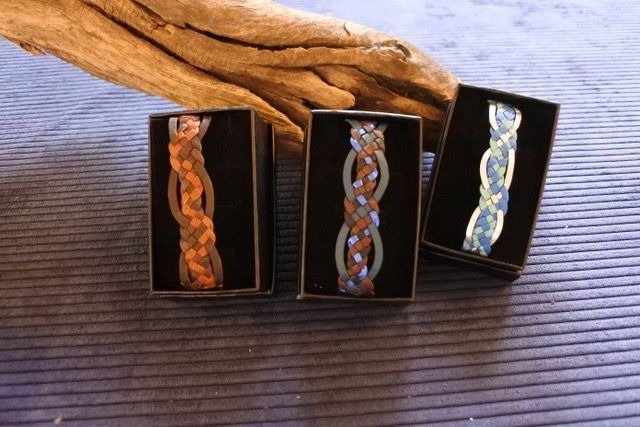 Three braided bracelets in small black boxes, in shades of brown, blue and white, with driftwood in the background.