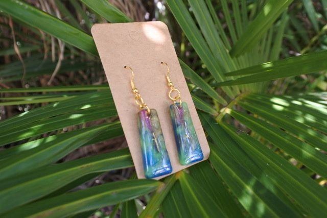Earrings with blue, green, and purple resin hang from gold hooks, displayed on a cardboard tag against green foliage.