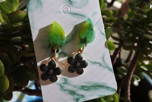 Earrings with green leaf-shaped tops and black flower pendants hanging on a marble-patterned card.