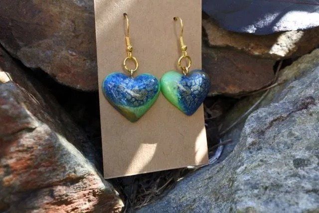 Heart-shaped earrings, blue and green, hanging from gold hooks, displayed on a brown card set against rocks.