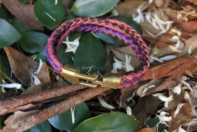 Braided leather bracelet, red and purple hues, gold clasp, on foliage.