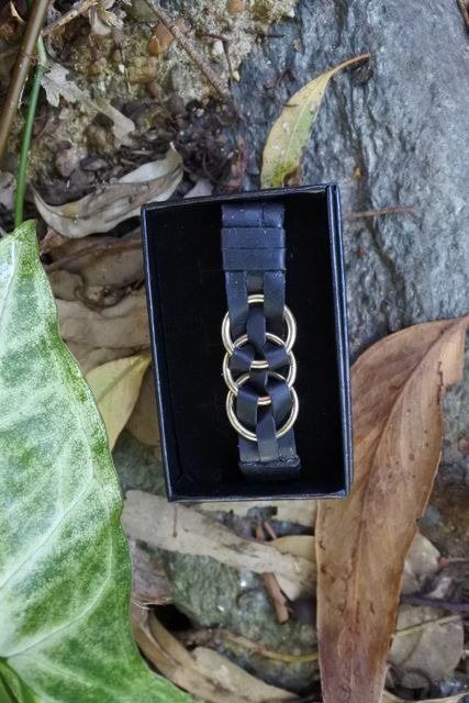 Black leather bracelet with gold rings in a black box, outdoors on rocks and leaves.