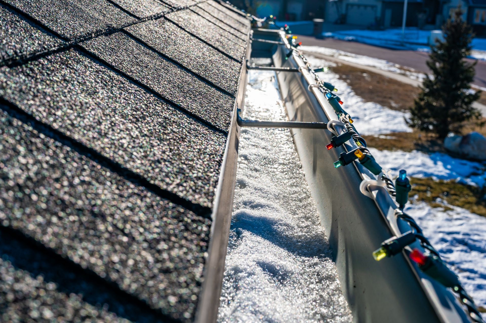 Close-up view of a snow-filled gutter on a roof, strung with colorful Christmas lights.