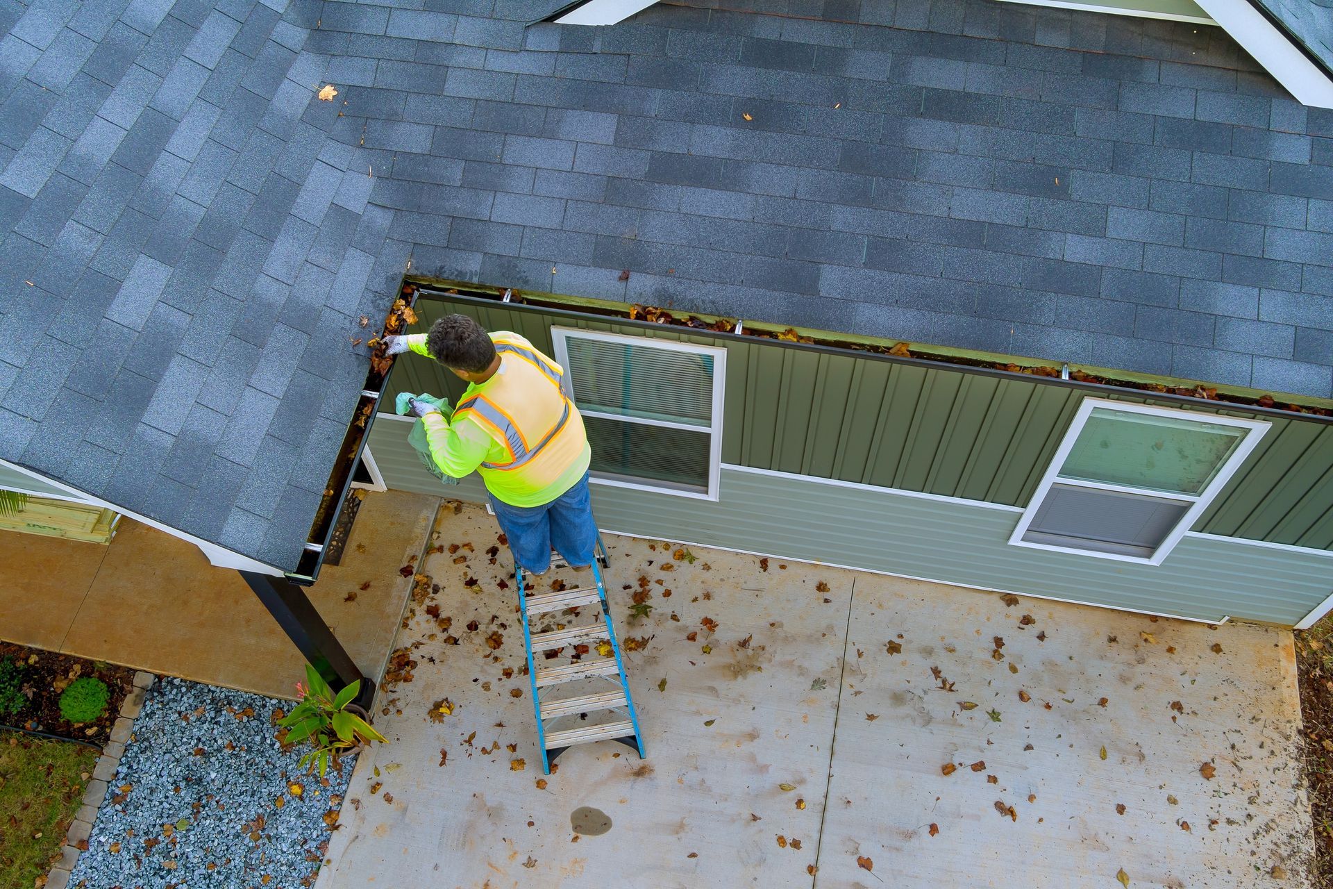 Person on a ladder cleaning gutters on a house. They are wearing a safety vest.