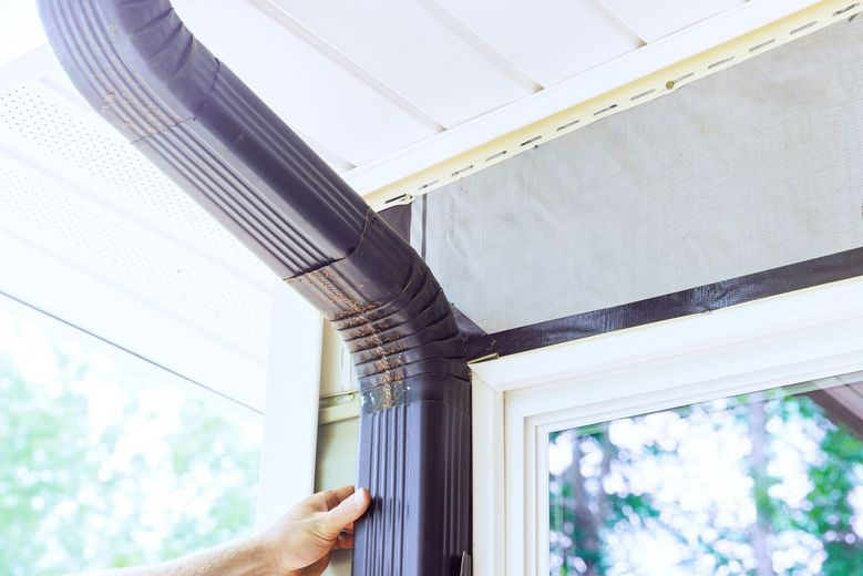 A hand installing a dark brown rain gutter on a house, next to a window.