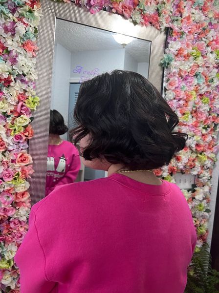 A woman in a pink sweater is standing in front of a mirror in front of a wall of flowers.