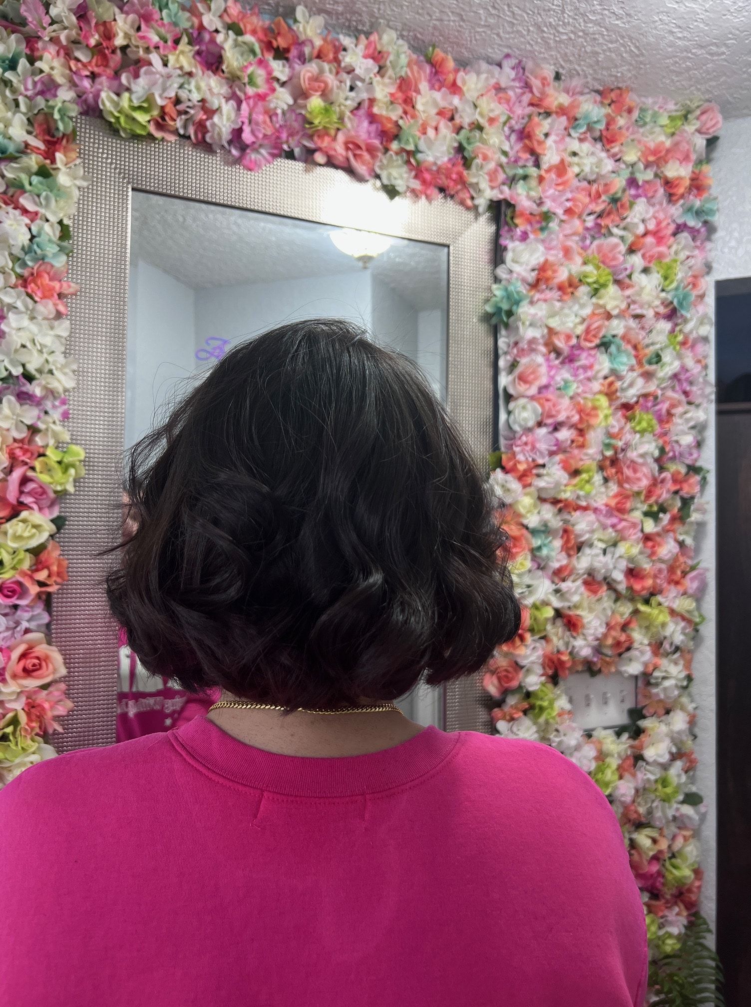 A woman in a pink shirt is standing in front of a wall of flowers.