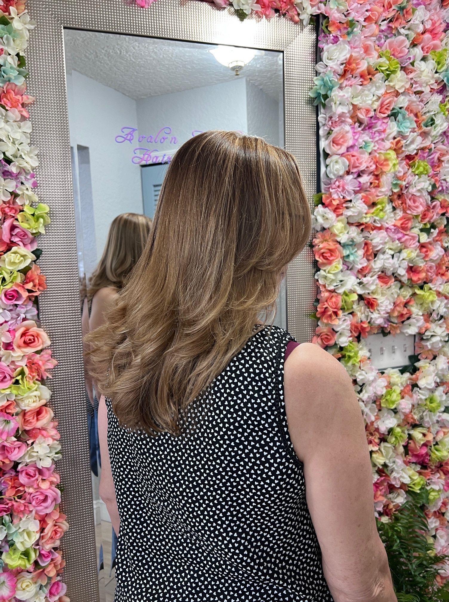 A woman is standing in front of a mirror in front of a wall of flowers.