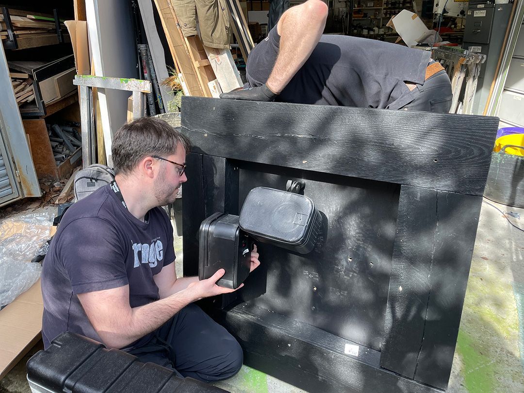 Two men are installing speakers into a large piece of wood.