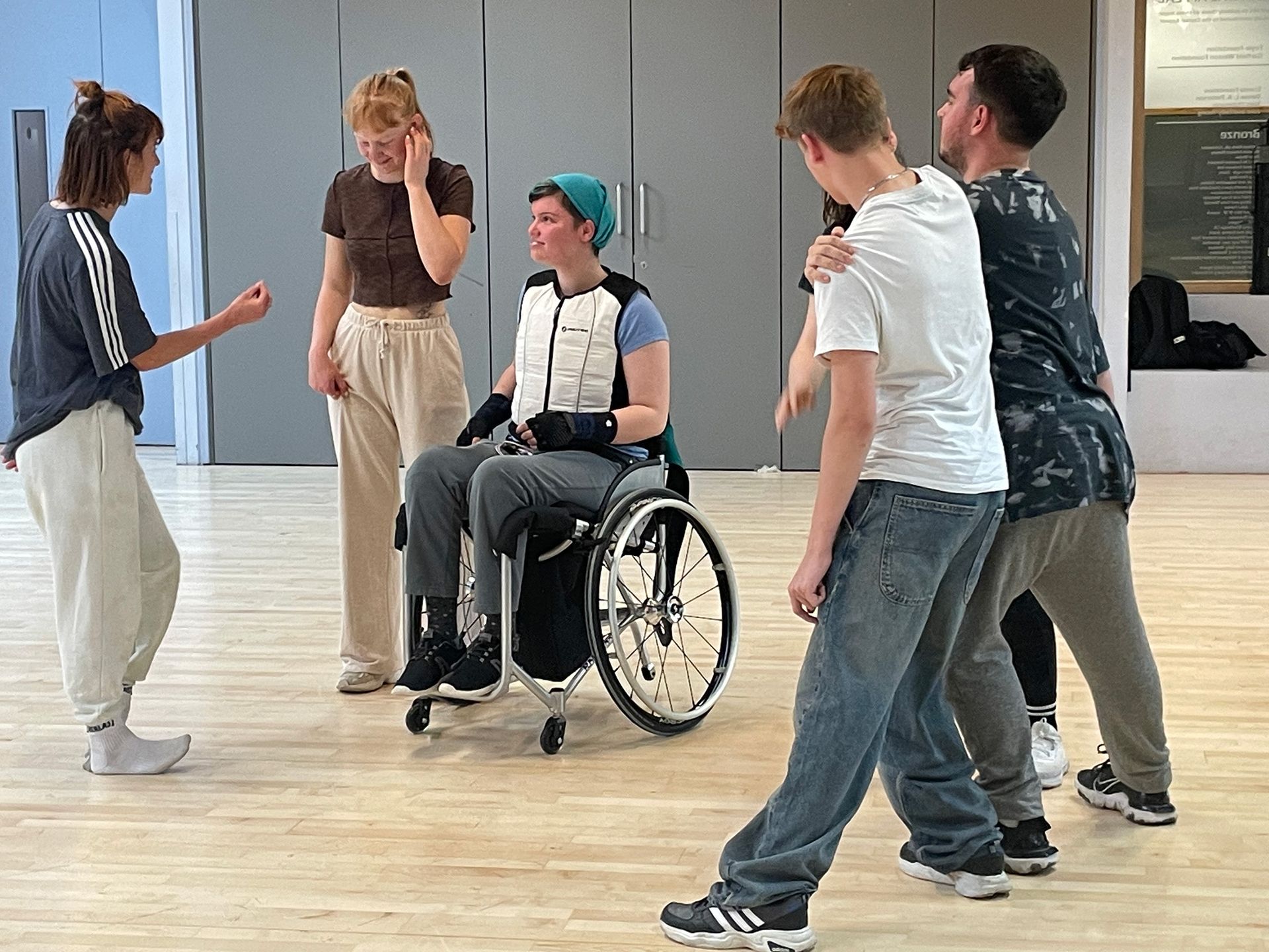 A group of people are standing around a woman in a wheelchair in a dance studio