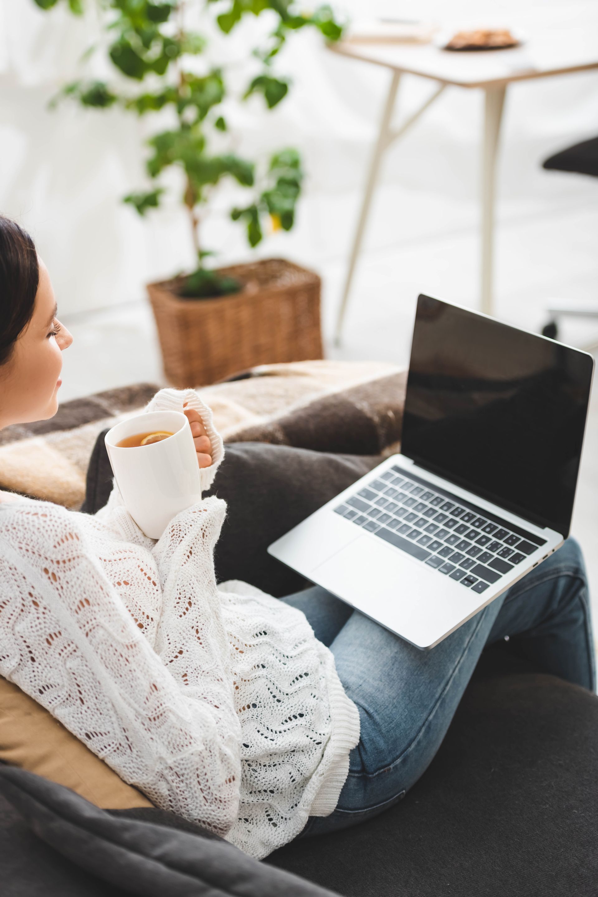 Woman relaxing on a couch, holding a mug and using a laptop; indoor setting with a plant.