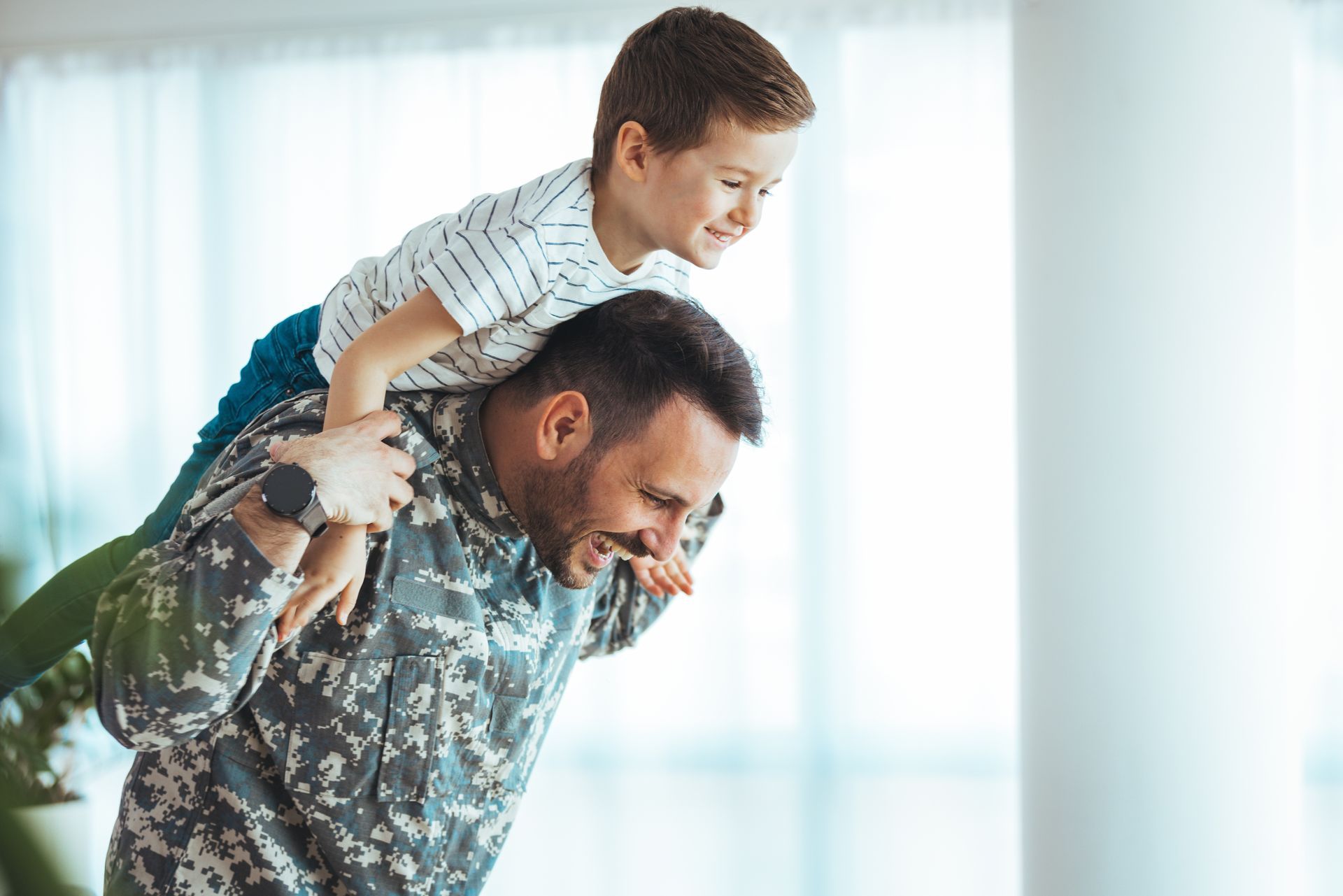 Man in camouflage carries a boy on his shoulders, both smiling in a bright room.