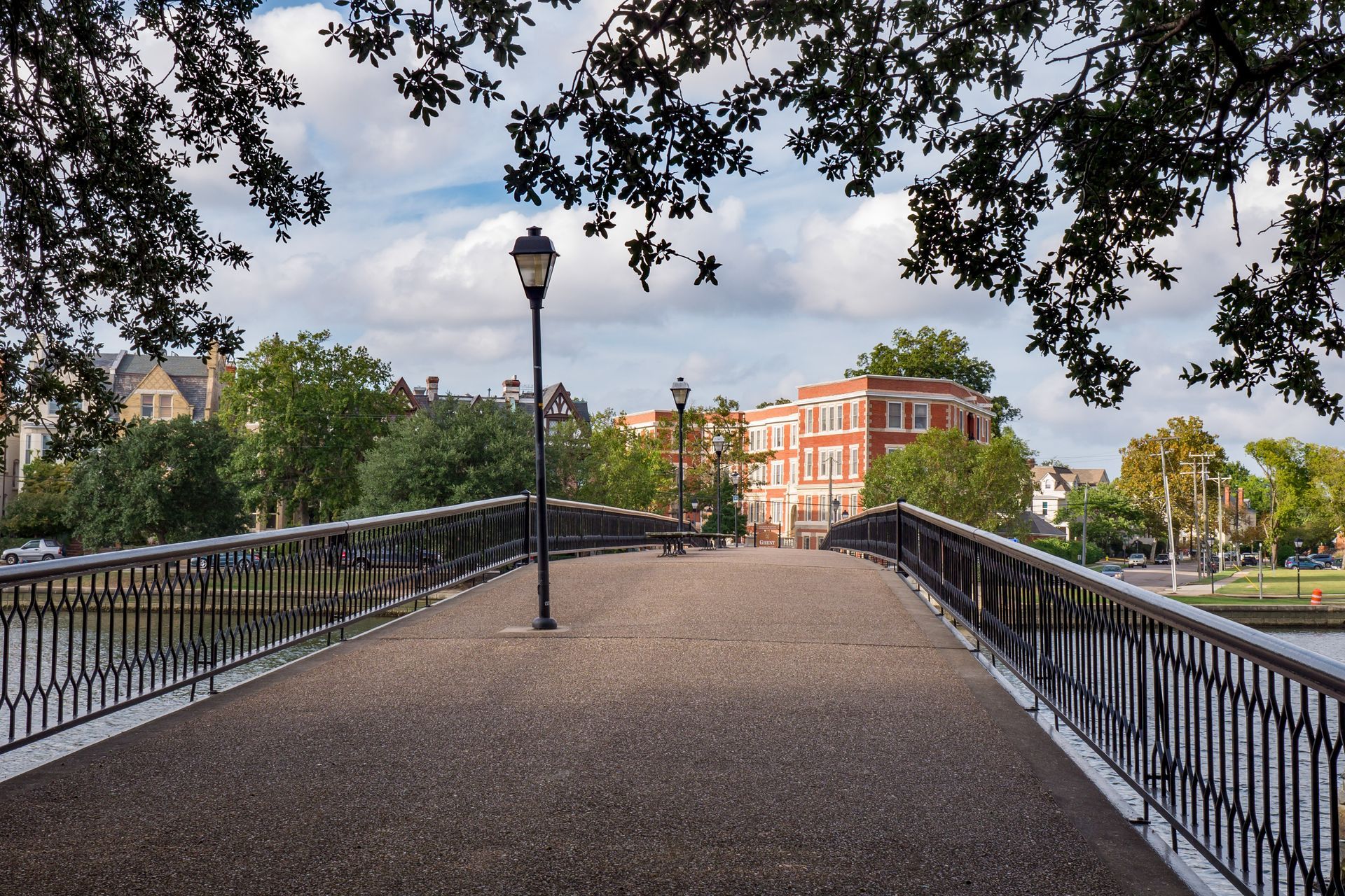 Bridge over water with a lamppost, trees, and buildings under a cloudy sky.