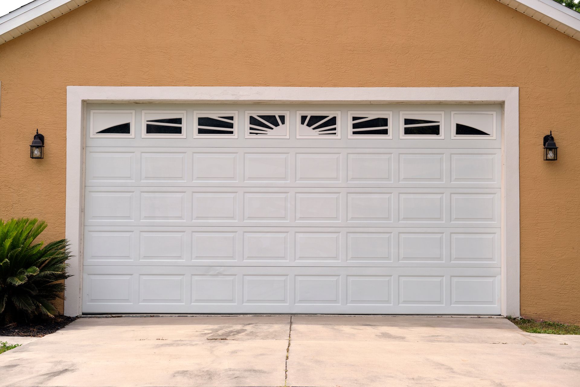 Beige house with white garage door, sunburst windows, black lights, and concrete drive.