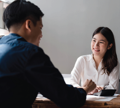 Two smiling people sit at a desk, discussing business. Woman in white shirt, man in blue.