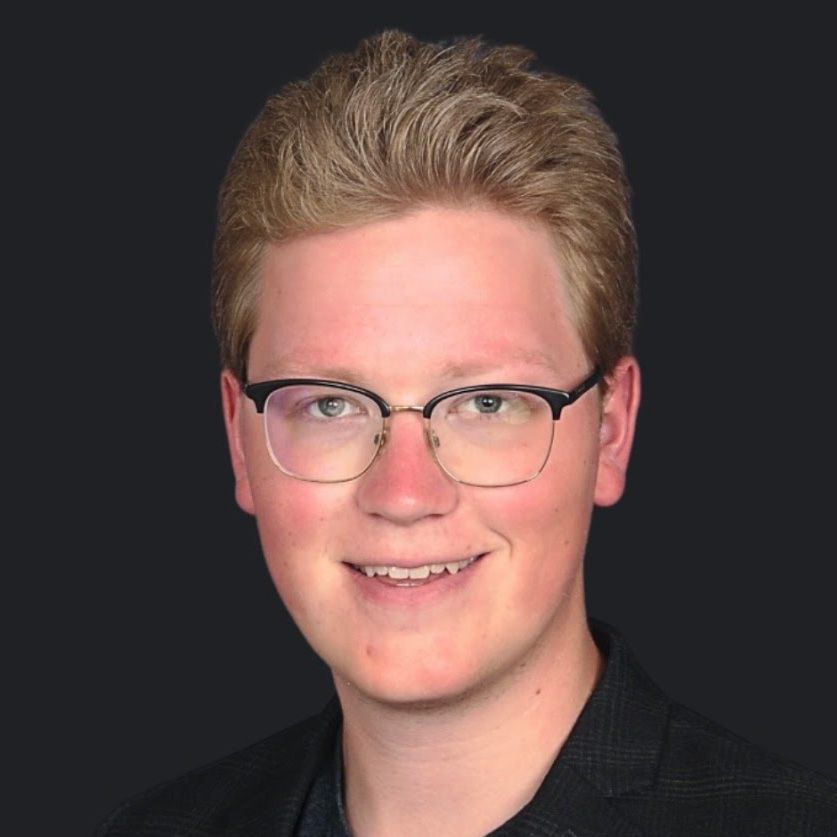 A young man wearing glasses and a black jacket is smiling for the camera.