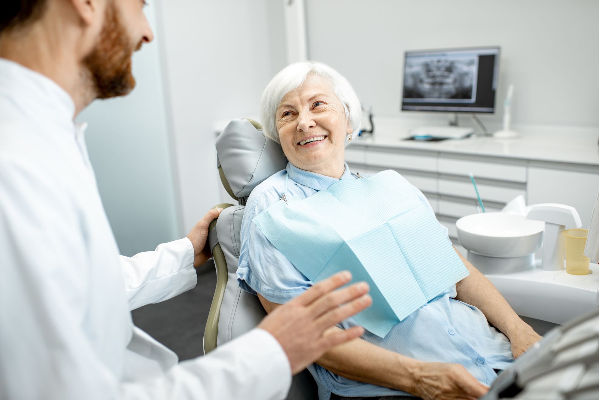 Dentist talking to smiling patient in a dental clinic, with dental chair and monitor in the background Dentist talking to smiling patient in a dental clinic, with dental chair and monitor in the background