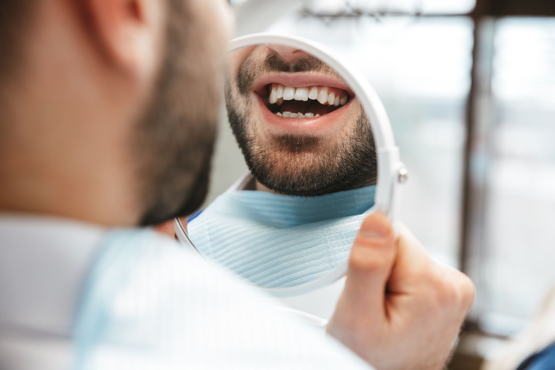 Person examining teeth in a handheld mirror, smiling with an open mouth in a bright room Person examining teeth in a handheld mirror, smiling with an open mouth in a bright room