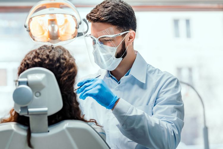 Dentist wearing mask and gloves examines a patient in a dental chair under bright exam light