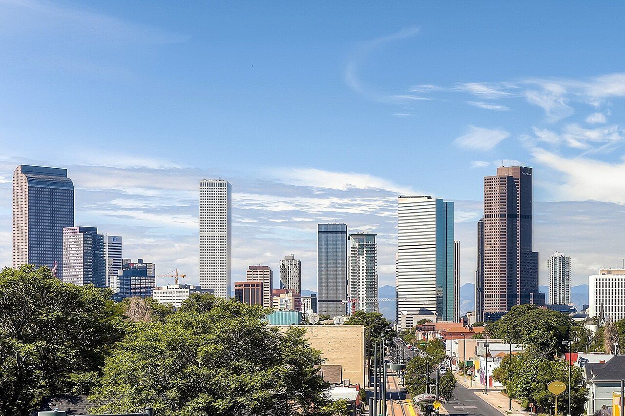 Skyline of tall modern buildings under a blue sky, seen above green trees.