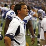 Football coach in white shirt with dark blue accents, looking up on the field, players in the background.