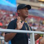 Man wearing a black cap and shirt, waving with open hands, at an outdoor stadium event.