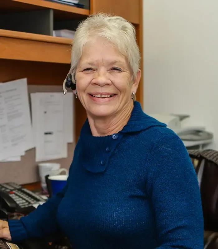 Woman with short white hair, smiling, wearing a blue sweater, sitting at a desk with papers and a phone.