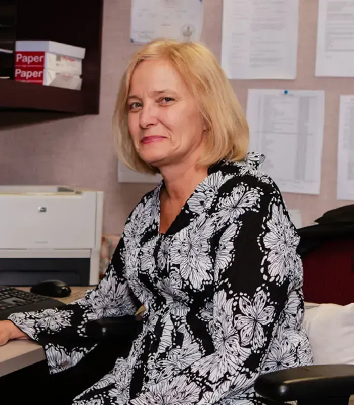 Woman with blonde hair wearing a black and white floral blouse, seated at a desk with a computer and printer.