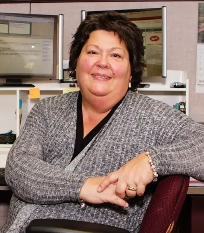 Woman smiling, seated in office setting, arms crossed, wearing gray cardigan.