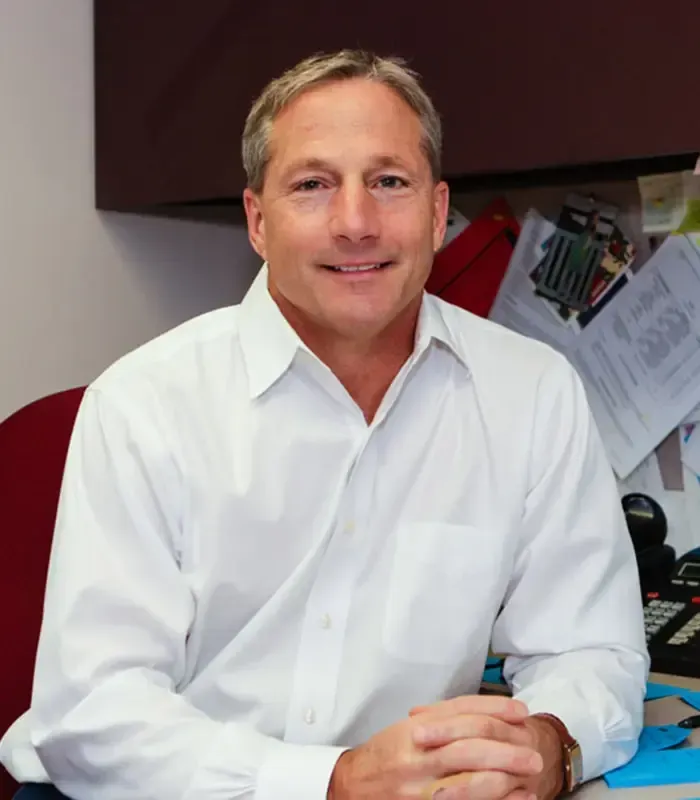 Man in white shirt smiling, sitting at desk with papers and phone, office setting.