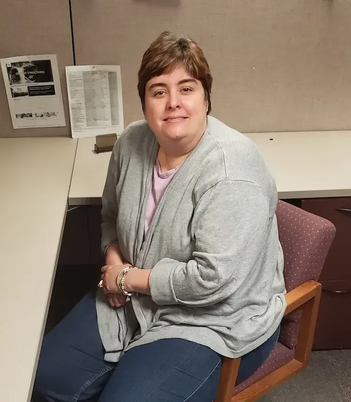 Woman seated at a desk, wearing a gray sweater, looking at the camera.