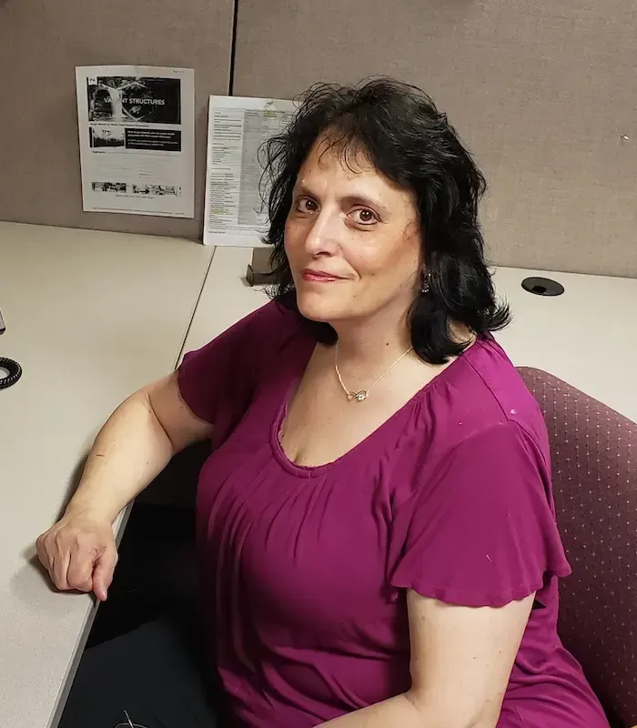 Woman in purple top seated at desk, smiling, with papers on the wall behind her.