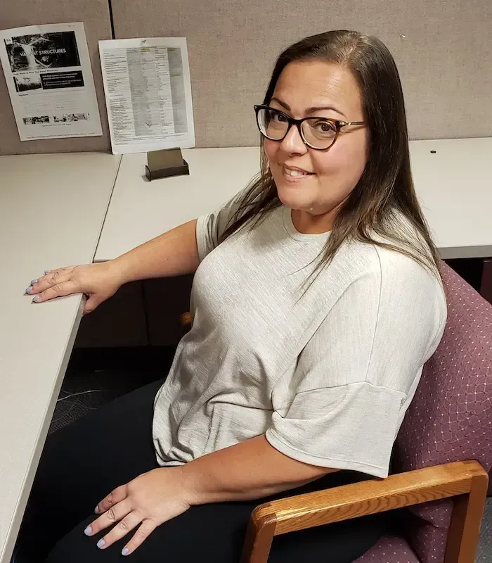 Woman in glasses, seated at a desk, smiles at the camera. She wears a light top and dark pants, in an office setting.