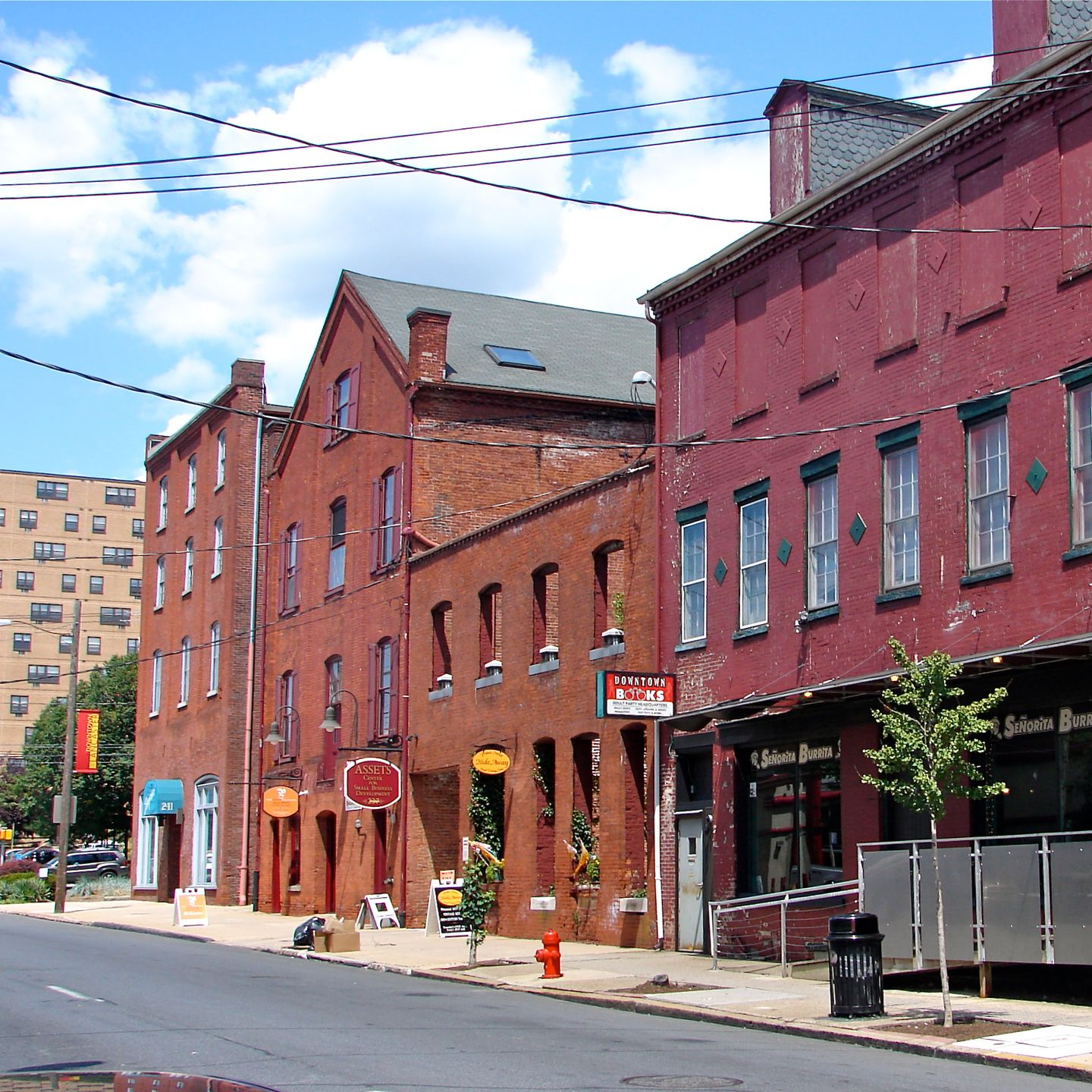 Buildings on a street, some red brick, with storefronts, overhead power lines, and blue sky.