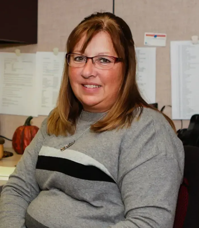 Woman wearing glasses and gray sweater smiles, seated indoors near a pumpkin and papers.