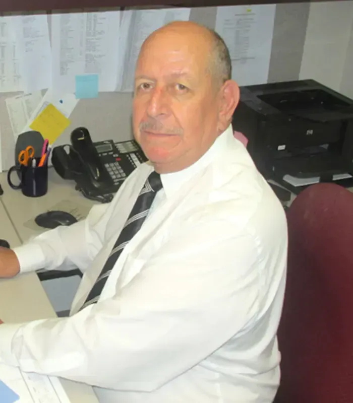 Man in white shirt and tie at a desk, looking at the camera. Office setting with phone and printer.