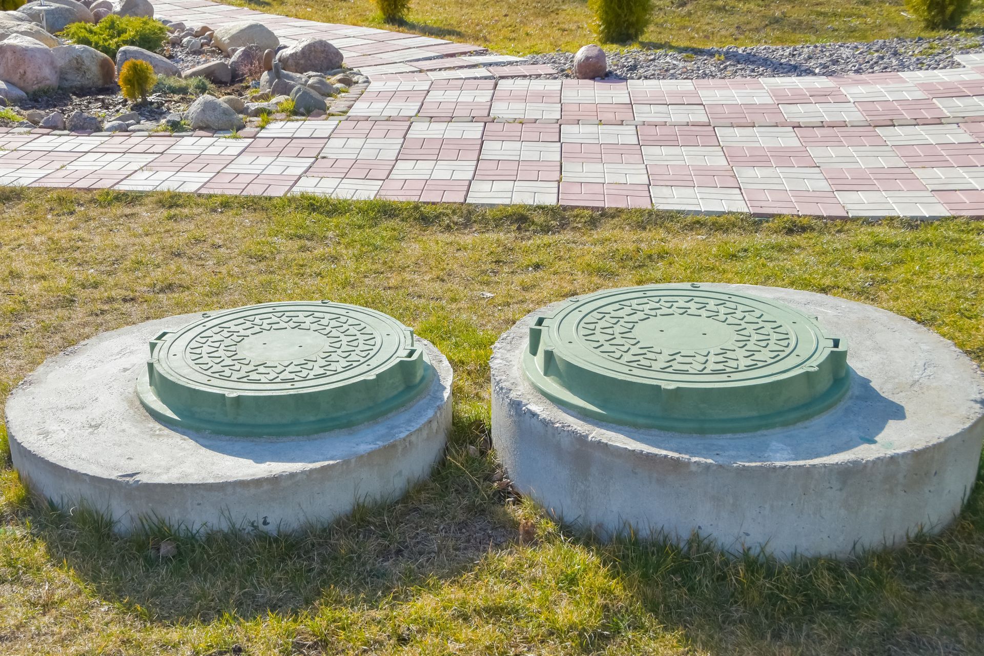 Two manholes of a septic system on the ground, next to a tiled road, below the sun.