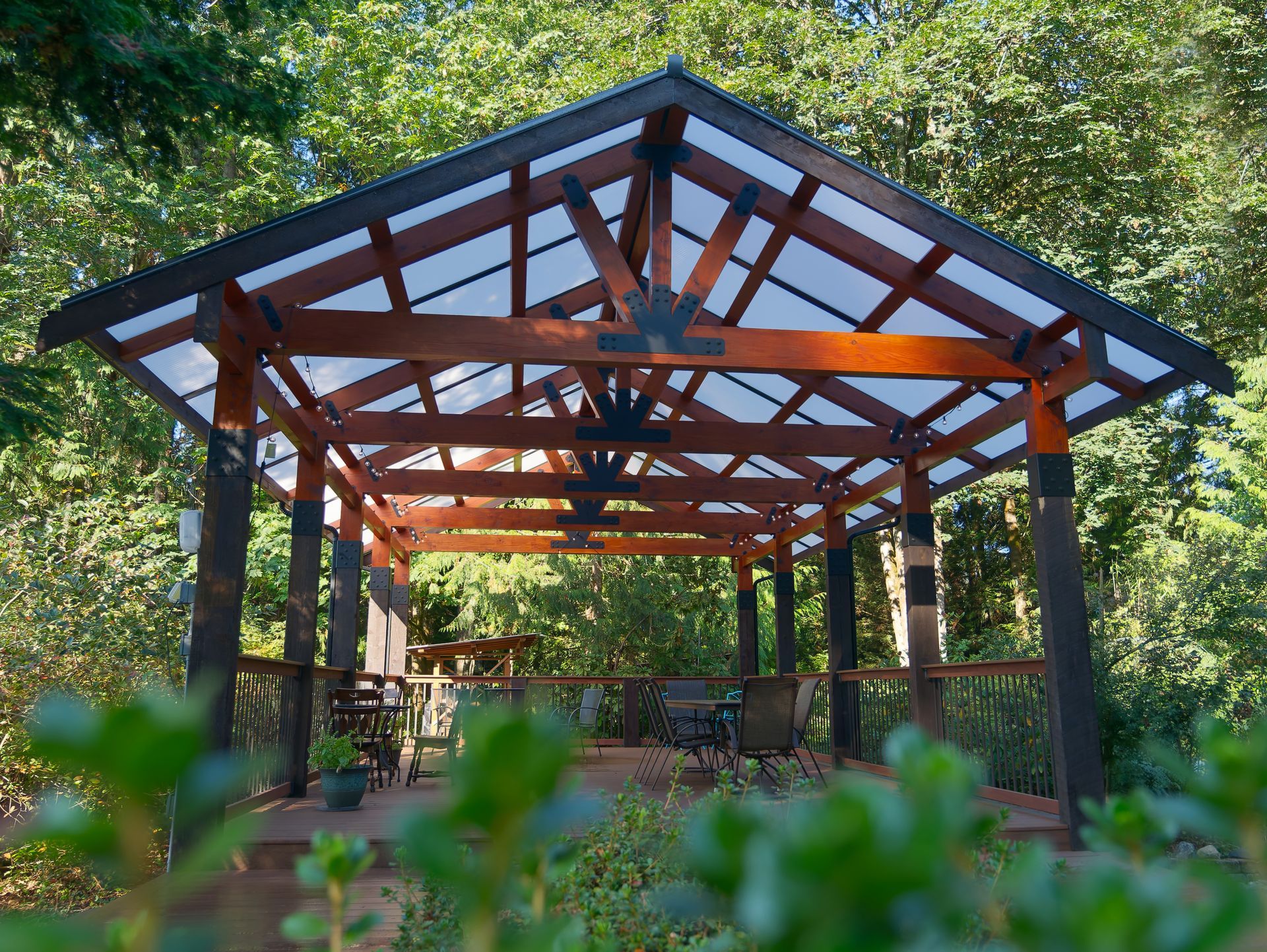 Wooden gazebo with translucent roof, in a wooded area. Interior table and chairs visible.