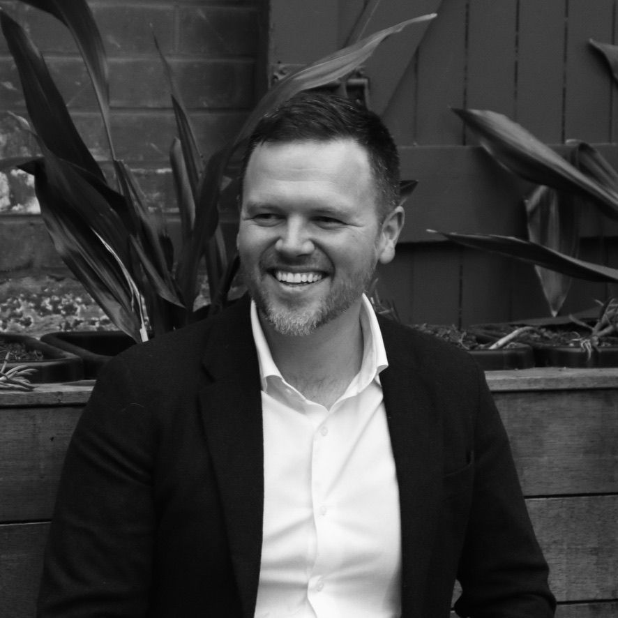 Smiling man in a dark suit and white shirt seated outdoors by large leafy plants