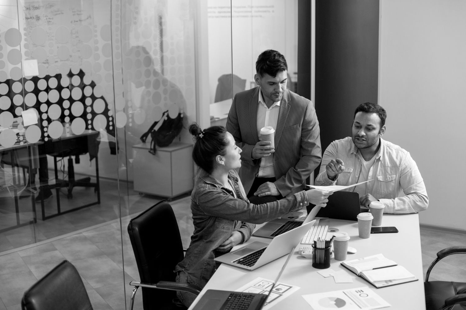 Coworkers in an office meeting, discussing papers around a table with laptops and coffee mugs.