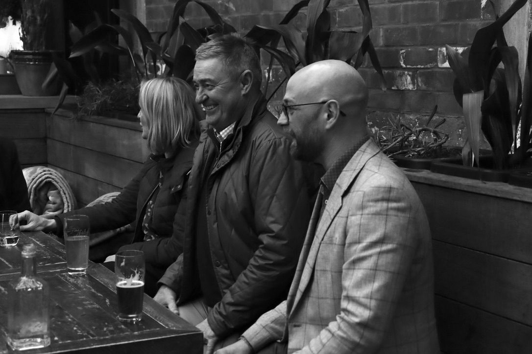 Three people seated at a bar in a black-and-white photo, chatting beside drinks.