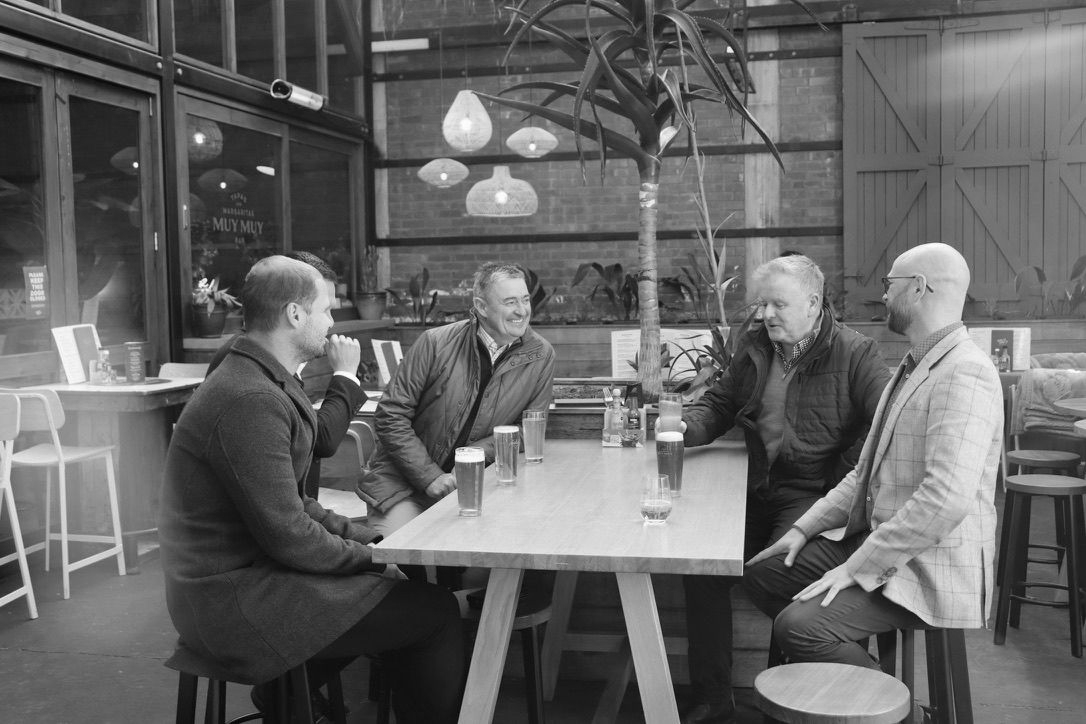 Four people sit talking around a table in a rustic café with hanging lights.