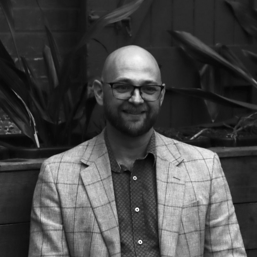 Smiling bald man in glasses wearing a plaid blazer, standing indoors against leafy plants.