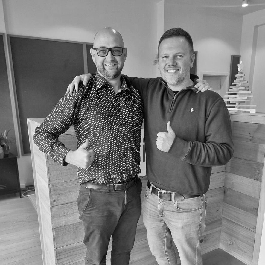 Two smiling men standing indoors, arm in arm, giving thumbs up in a lobby