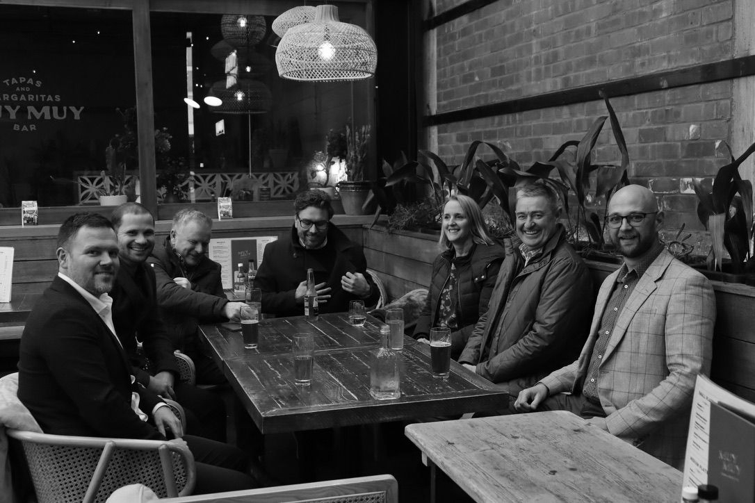 Six people seated around a wooden table in a cozy indoor restaurant or cafe, smiling at the camera.