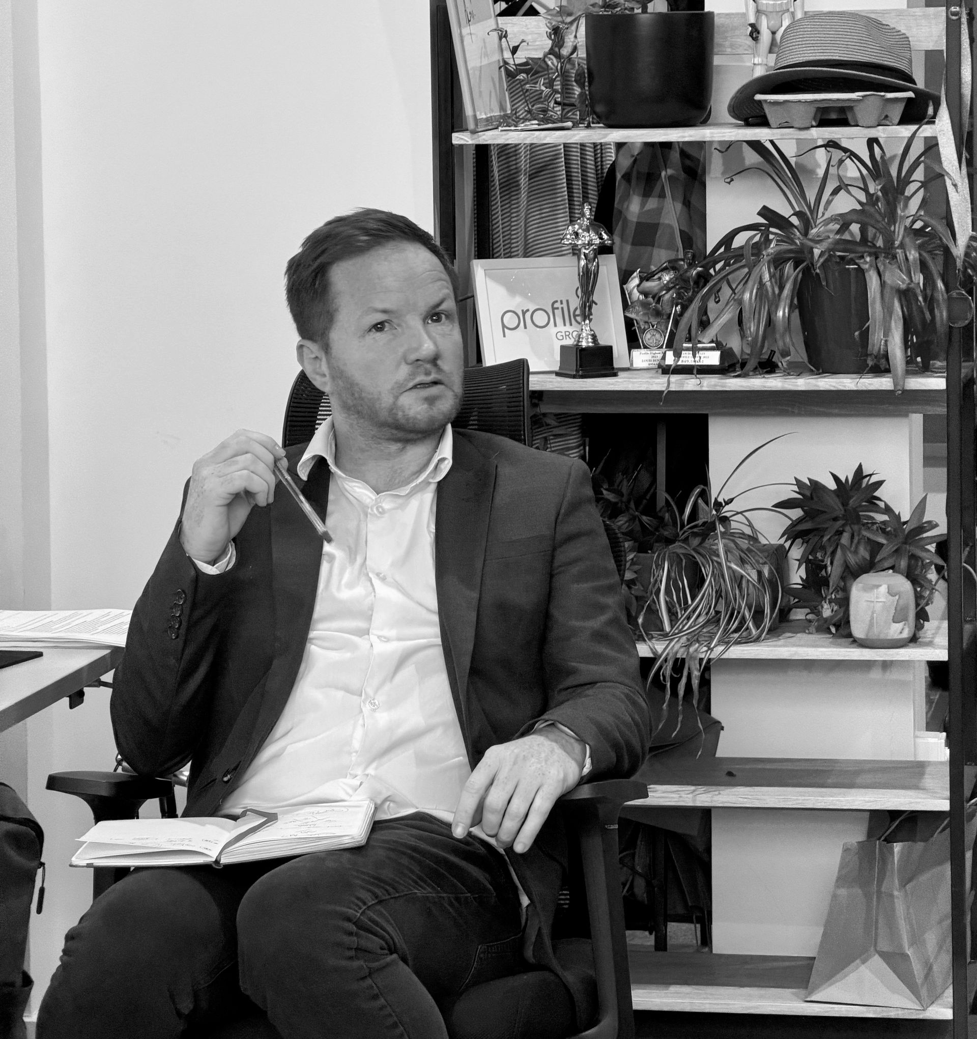 Man in a blazer seated in an office, holding glasses, with plants and shelves behind him.