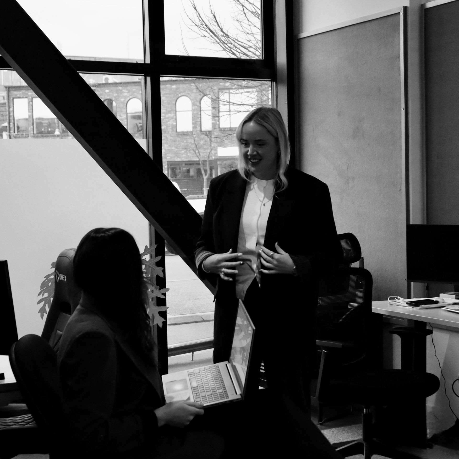 Two people talking in a bright office, one standing and smiling near a desk, one seated by a window